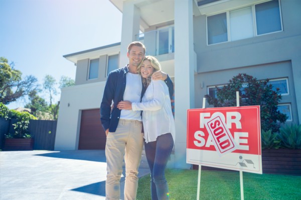 Buying a property for a short-term rental can prove lucrative. Here we see a couple standing next to a sold sign after buying an investment property in 2024. They are smiling at the camera.  