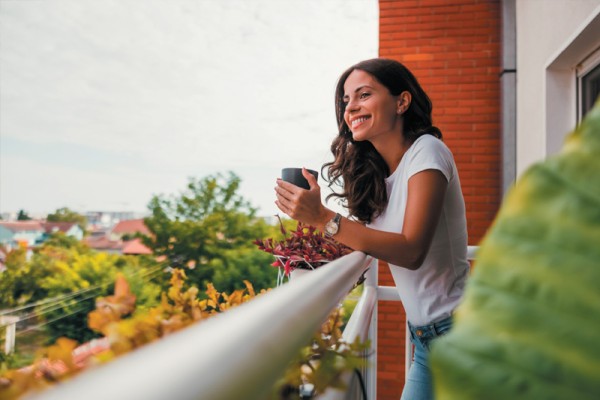 The Melbourne property market in 2024 has seen more families opting for apartments. Here we see a young woman on a balcony enjoying the view from her apartment. She is drinking a from a mug and smiling. 