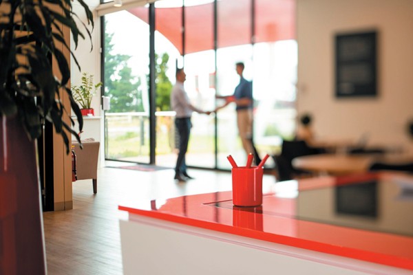 Prioritising buyer support and education is a vital part of your property development sales strategy. Image of two men shaking hands at a sales office. They are blurred in the background. In the foreground is a reception desk with red top. 